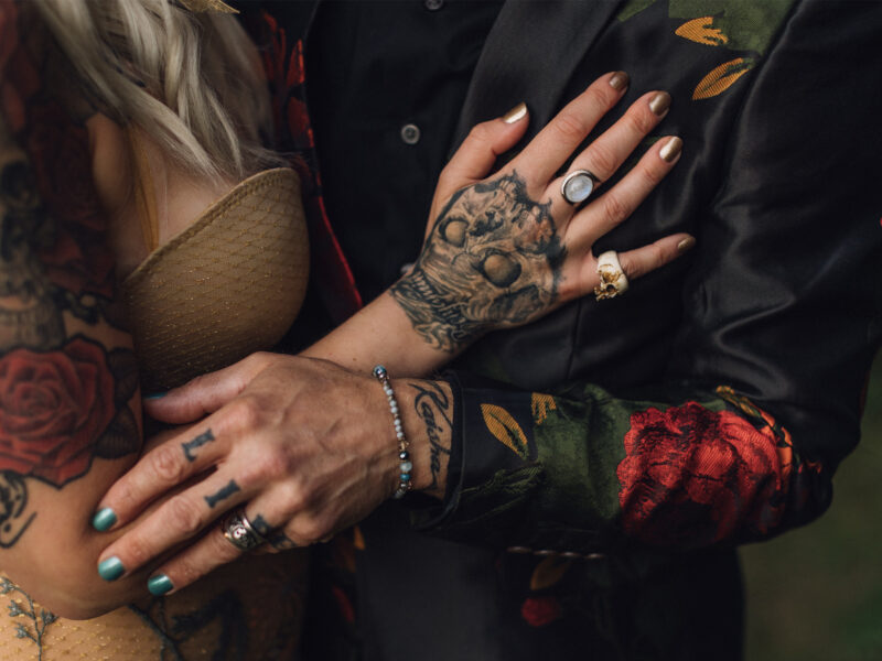 A tattoo artist couple embraces after getting married. A close up of their tattooed hands with their wedding rings.